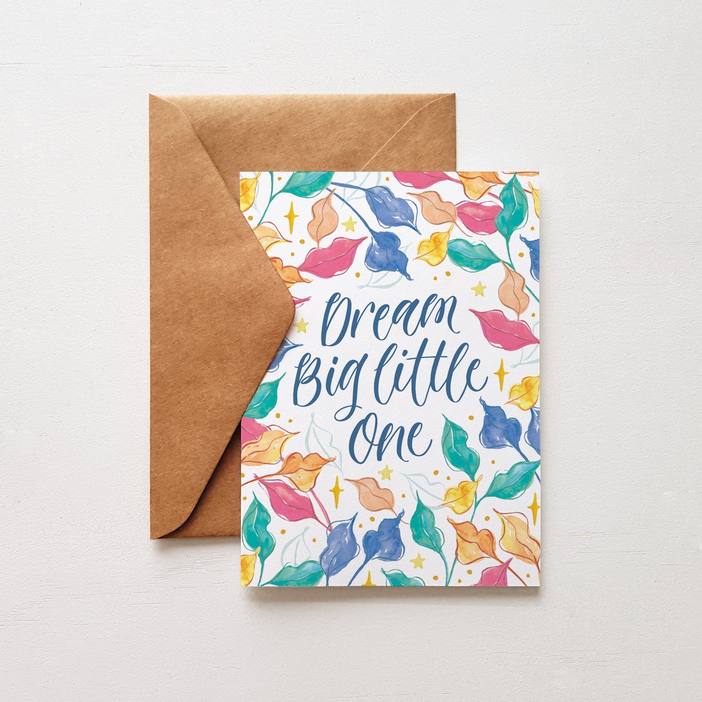 Greeting card with colorful leaf design and 'Dream Big little One' text, accompanied by a brown envelope on a light grey background.