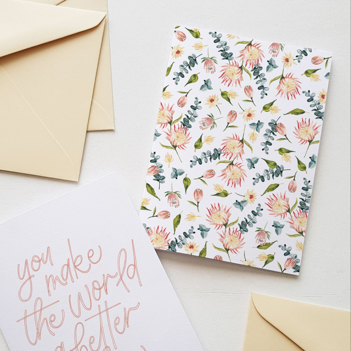 Greeting card with a soft pink protea flower pattern, displayed surrounded by cream coloured envelopes.