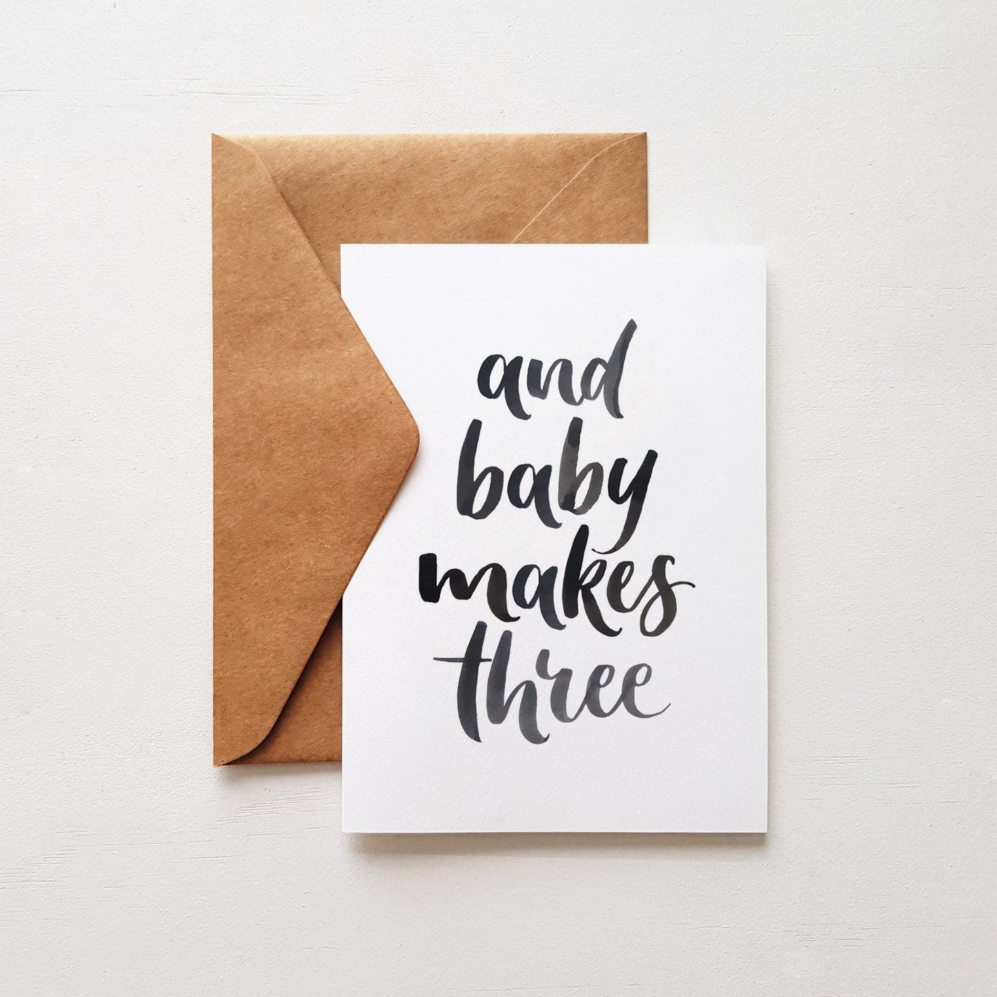 Greeting card with black hand lettering 'and baby makes three' with a brown envelope