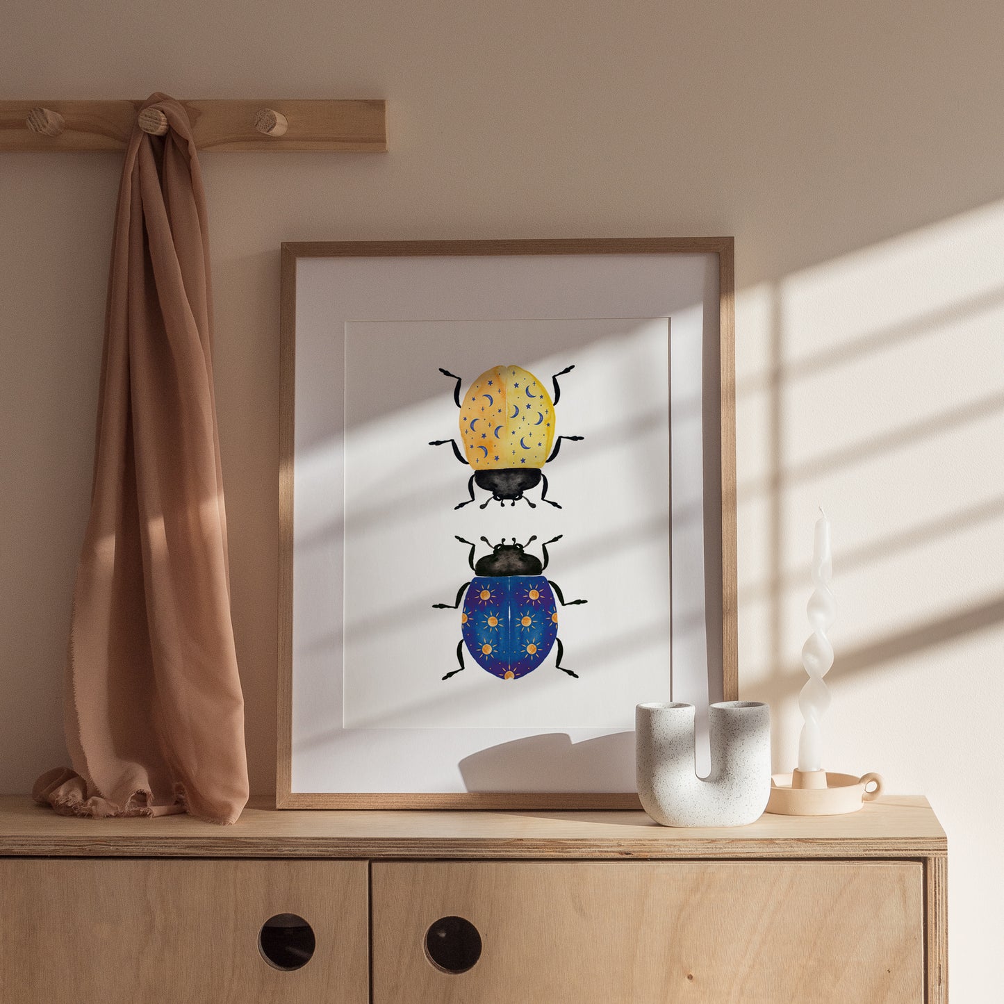 wooden framed domino lady bug sunny days and starry nights artwork on top of a cabinet. a brown throw hangs on a wooden rack in the top left corner. ceramic pieces sit to the bottom right of the wooden frame