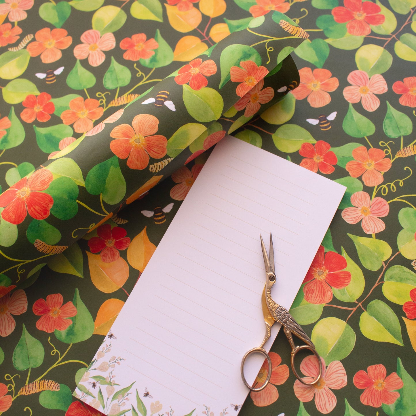 rolled wrapping paper with illustrated garden poppies. White notepad with lines, golden scissors sit onto of the lined note pad