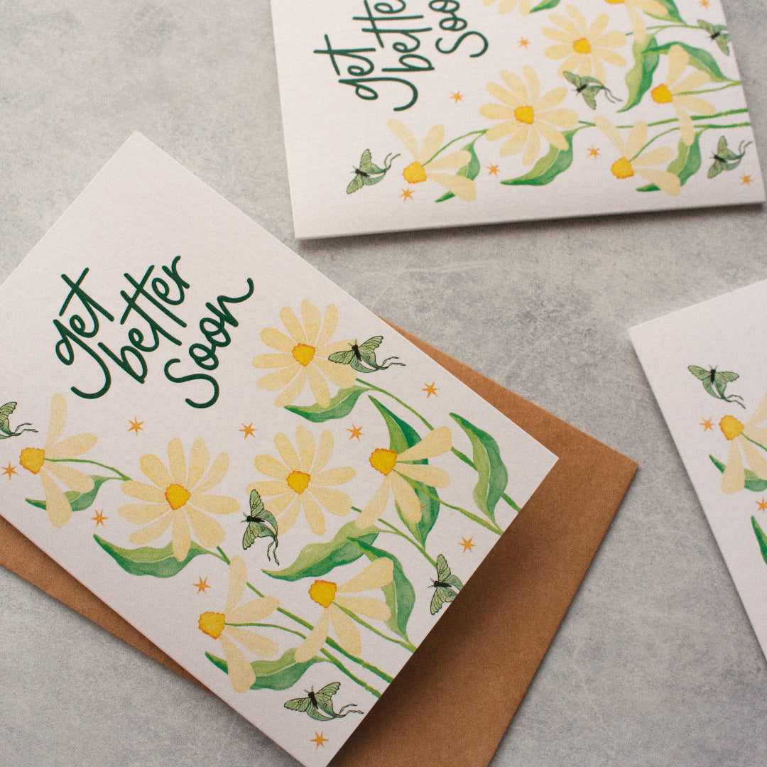Get well soon greeting cards featuring cream daisies, teal luna moths, and a kraft envelope. The card displays 'Get Better Soon' in deep green hand lettering, offering a heartfelt message of comfort and encouragement.