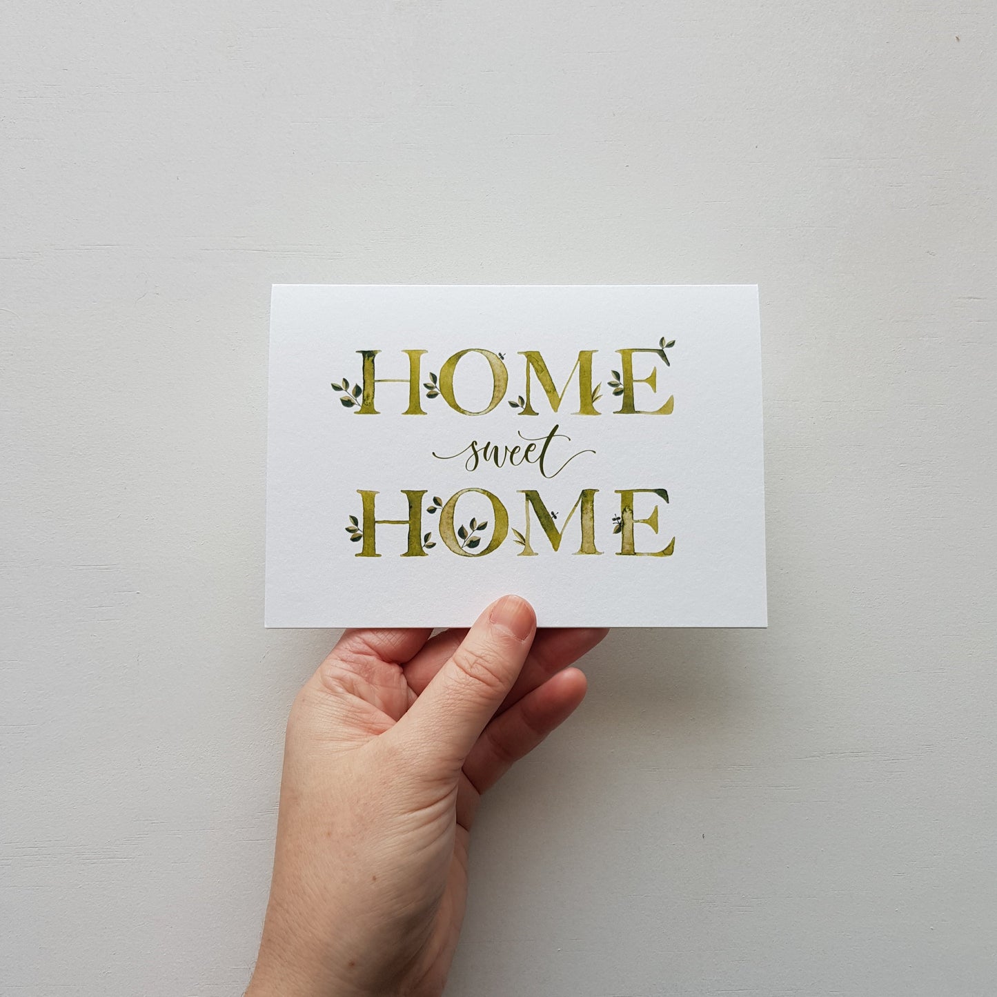 A hand holding a white square card with the phrase 'HOME sweet HOME' printed in gold decorative lettering against a plain light background.