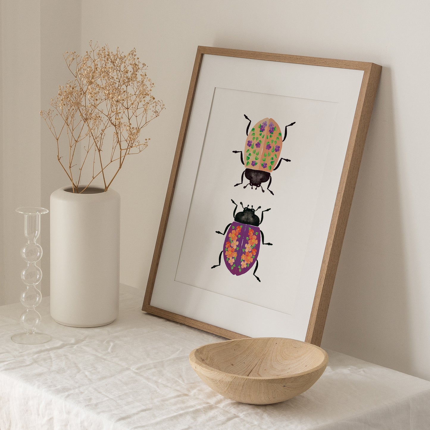 two lady bugs face each other in a wooden frame. One lady bug is upside down with a peachy orange back with vines and purple flowers. The other lady bug has a deep purple back with peachy orange poppy flowers. The frame sits on a table with a white table cloth. A white vase with dried flowers is positioned to the left of the frame as well as a clear slim decorative vase. An empty wooden bowl sits near the bottom right of the framed artwork.