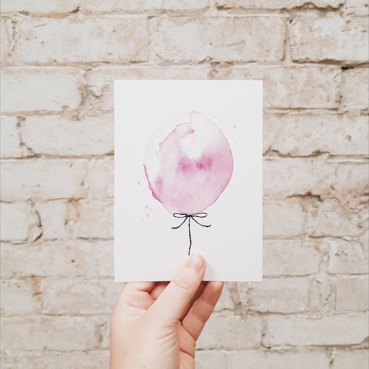 Hand holding a pink balloon greeting card against a brick wall