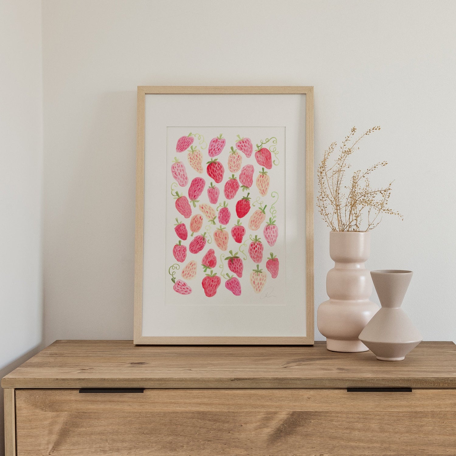 Framed artwork with pink strawberry design on a wooden surface with vases.