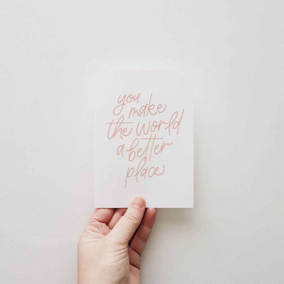 Hand holding greeting card with pink lettering 'you make the world a better place' on a white wall.