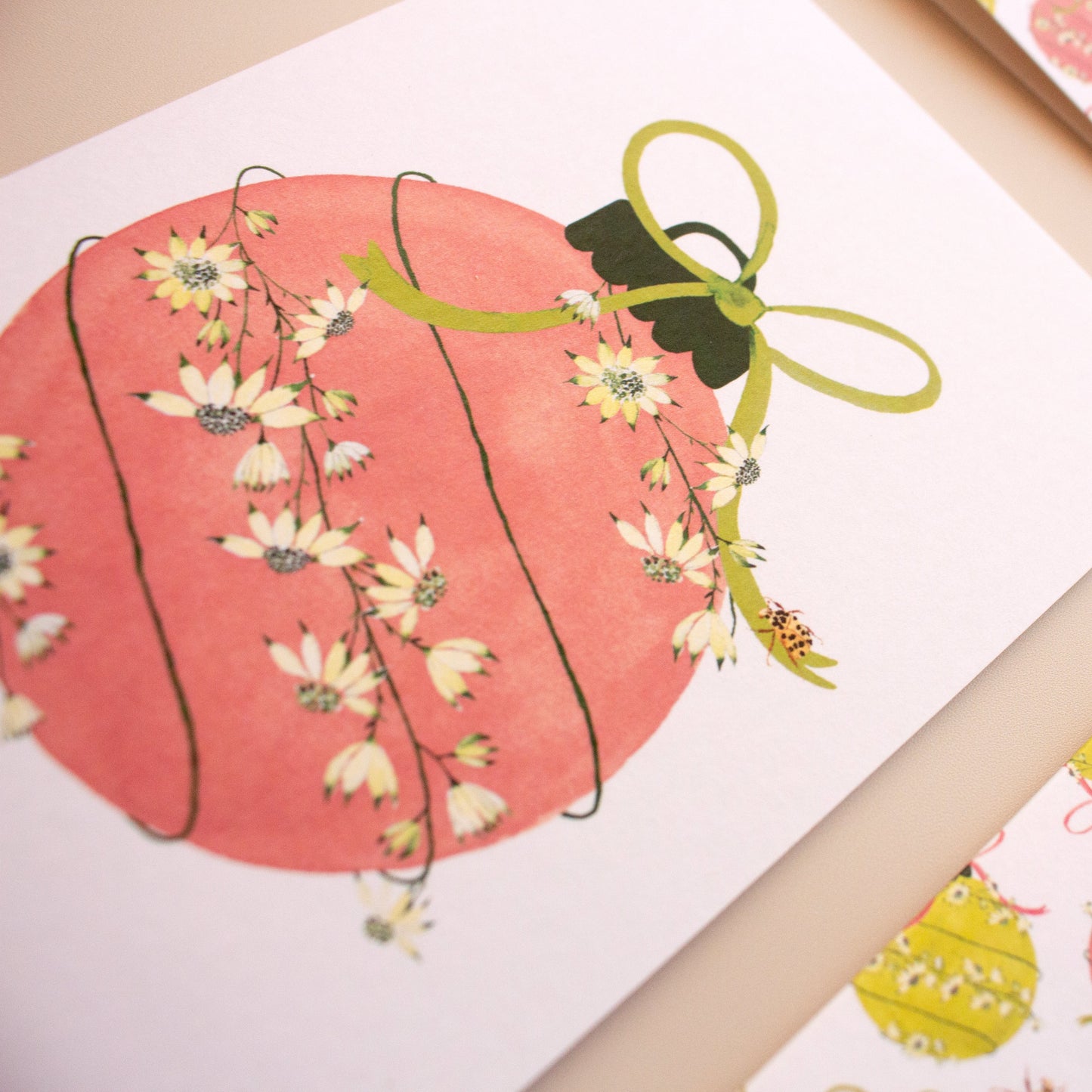 A close-up of printed cardstock featuring a watercolor-style pink ornament with white flowers and a green ribbon, alongside other patterned cards in pink, yellow, and green on a beige surface.