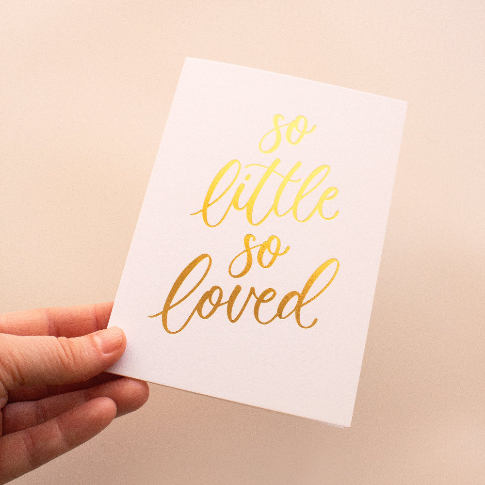 Hand holding a white card with 'so little so loved' in gold text on a beige background