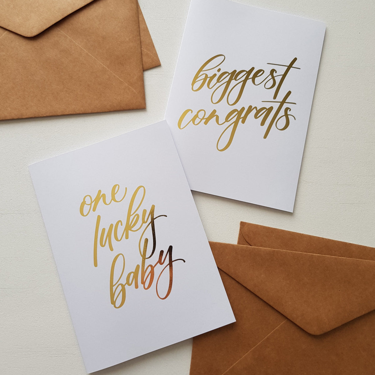 Two white greeting cards with gold foil script text, one reading 'one lucky baby' and the other 'biggest congrats'.
