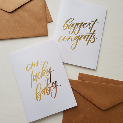 Two white greeting cards with gold foil script text, one reading 'one lucky baby' and the other 'biggest congrats'.