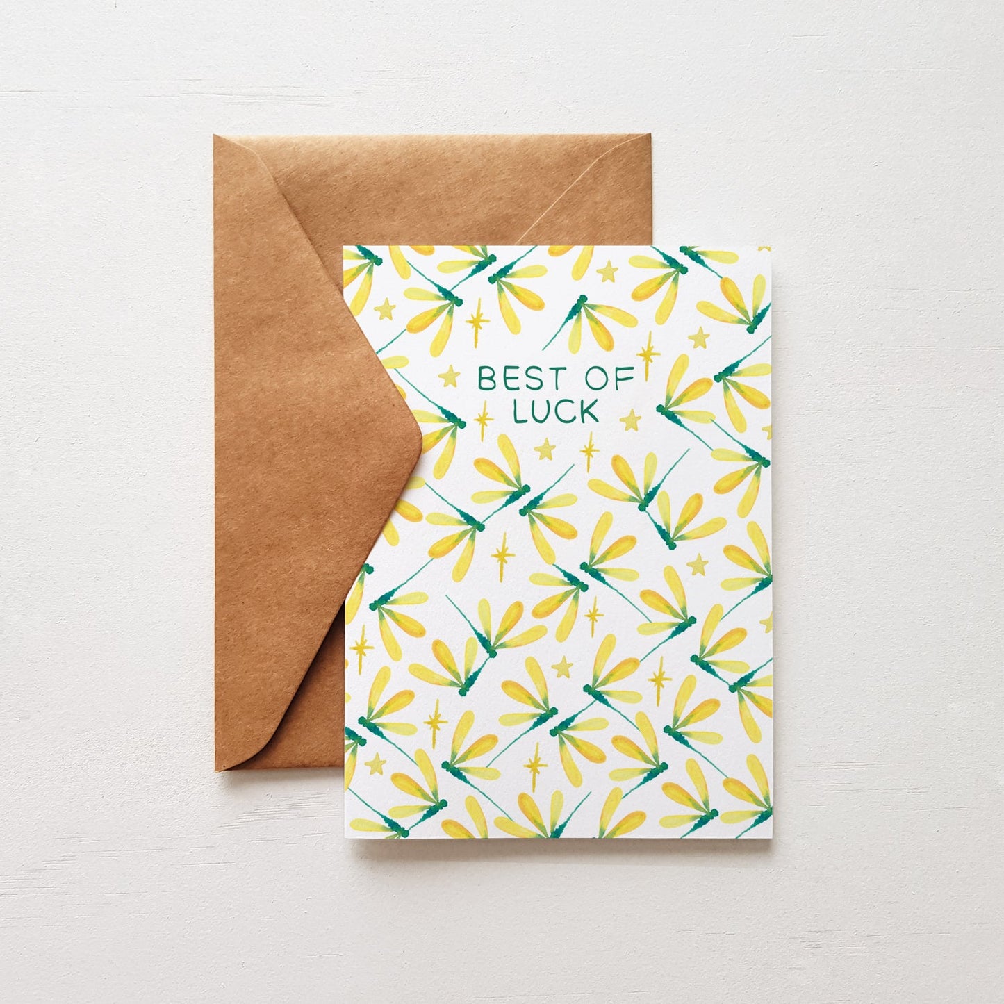 Greeting card with 'Best of Luck' text and dragonfly pattern, accompanied by a brown envelope on a light gray background.