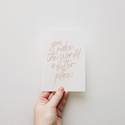 Hand holding greeting card with pink lettering 'you make the world a better place' on a white wall.