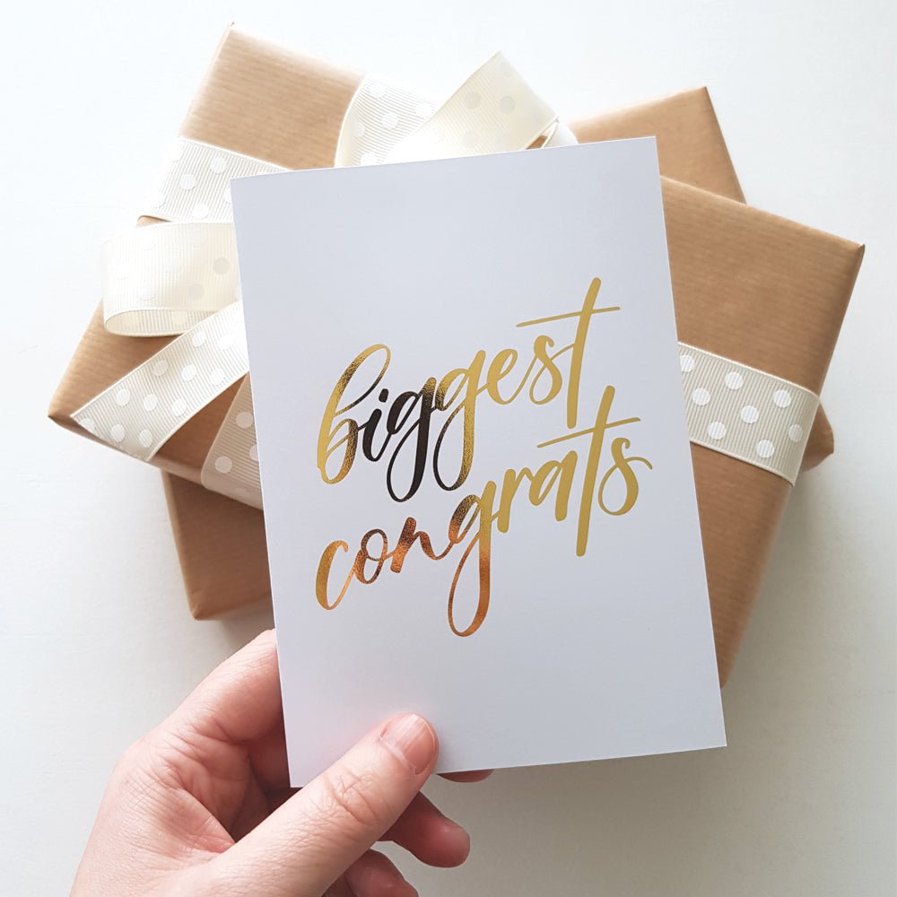 Hand holding a gold foil greeting card with text 'biggest congrats' next to warpped brown presents with cream ribbon