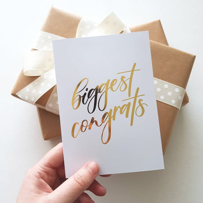 Hand holding a gold foil greeting card with text 'biggest congrats' next to warpped brown presents with cream ribbon