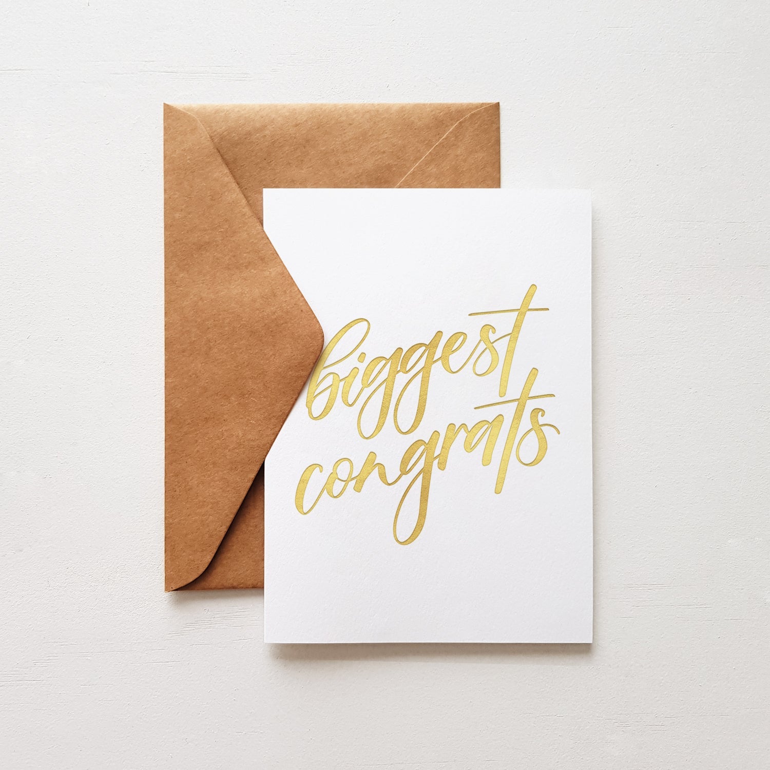 Greeting card with 'Biggest congrats' written in gold and a brown envelope on a white background