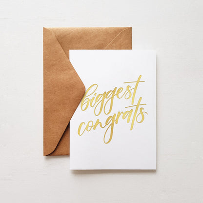 Greeting card with 'Biggest congrats' written in gold and a brown envelope on a white background