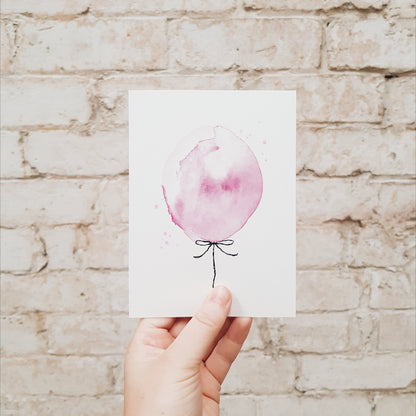 Hand holding a pink balloon greeting card against a brick wall