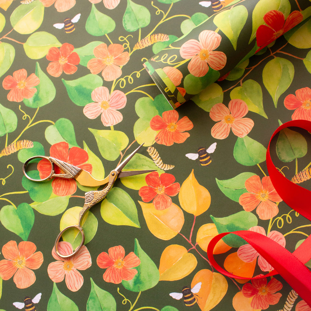 gold bird scissors on a hand painted foliage and floral design. Top right corner sits a roll of wrapping paper and bottom right hand corner a red ribbon