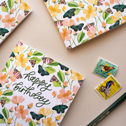 Three floral and butterfly-themed 'Happy Birthday' greeting cards arranged on a beige surface, with two small vintage-style stamps and a green pencil nearby.