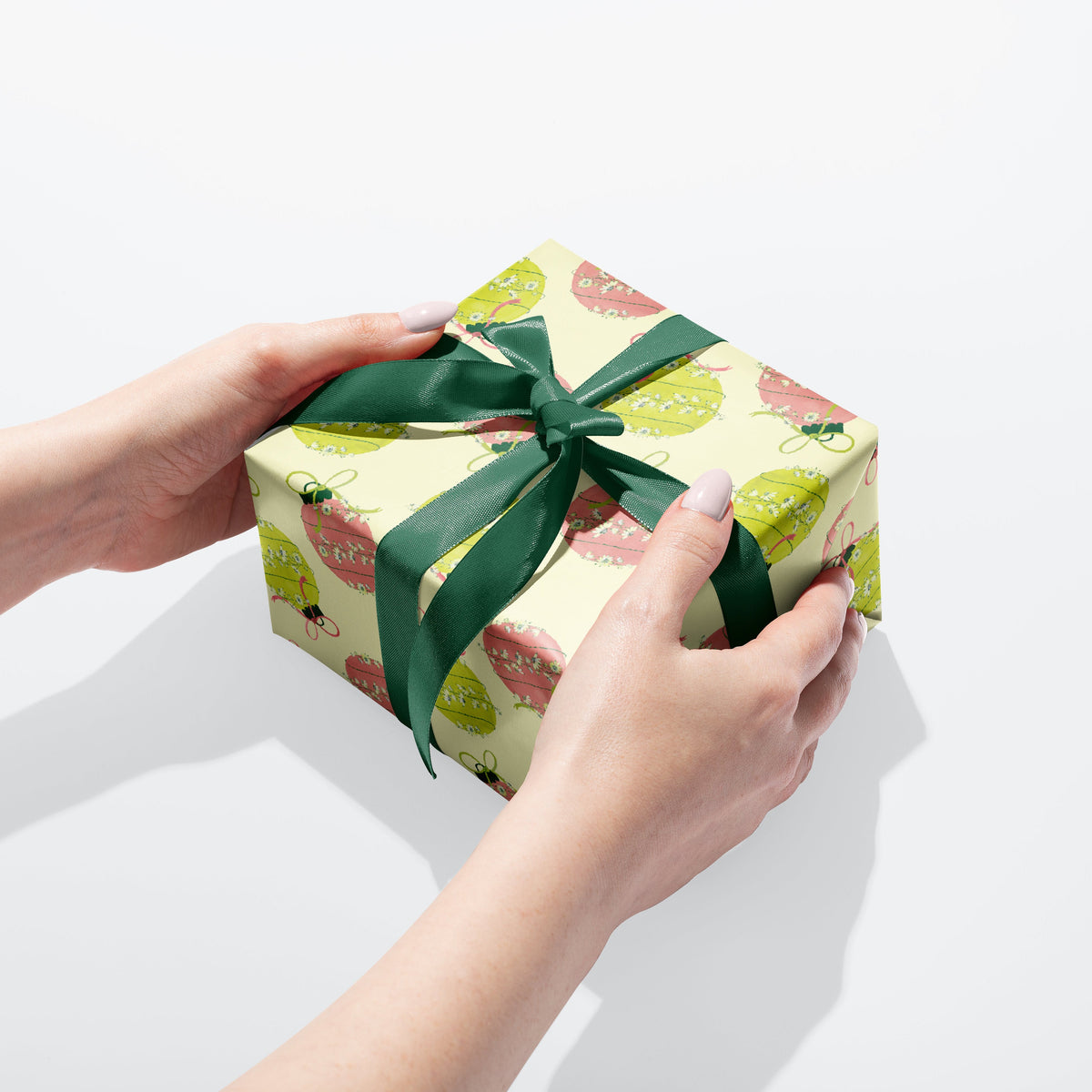 A pair of hands holding a square gift box wrapped in light yellow wrapping paper with a pink and light green Christmas and tied with a dark green ribbon, on a clean white background.