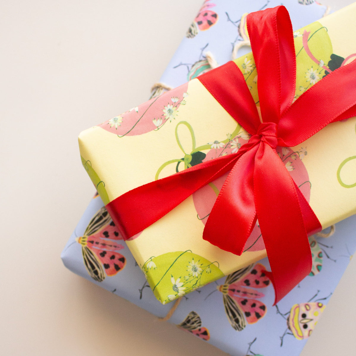 Two wrapped gift boxes stacked diagonally on a light beige background. The top box is wrapped in pale yellow paper with a large red satin bow, while the box beneath it is wrapped in light blue paper with a colorful bug pattern.