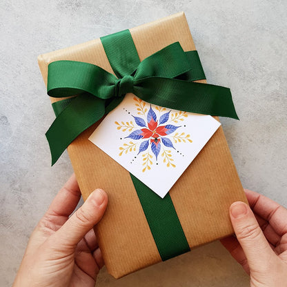 A wrapped gift box in brown kraft paper with a large green ribbon bow and a decorative white tag featuring a colorful floral snowflake design. Two hands are holding the box against a light gray textured background.