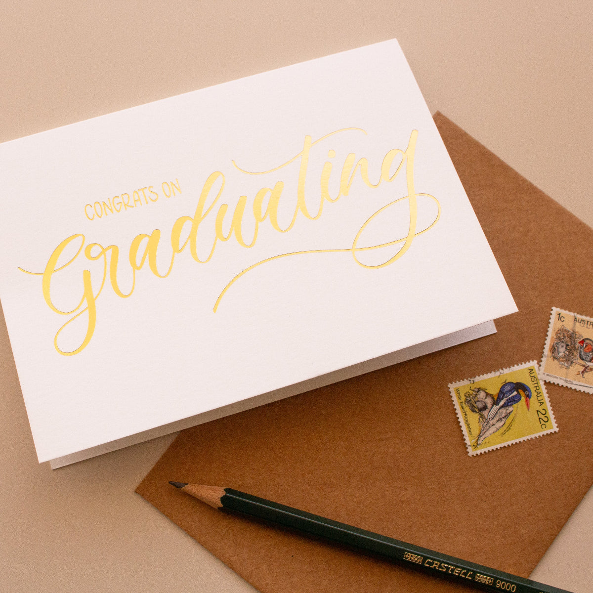 Graduation card with gold text on a brown envelope with stamps and a pencil.