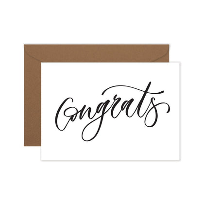 A folded white greeting card with the word 'Congrats' written in a elegant, hand lettered script, and a brown kraft envelope.