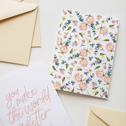 Greeting card with a soft pink protea flower pattern, displayed surrounded by cream coloured envelopes.