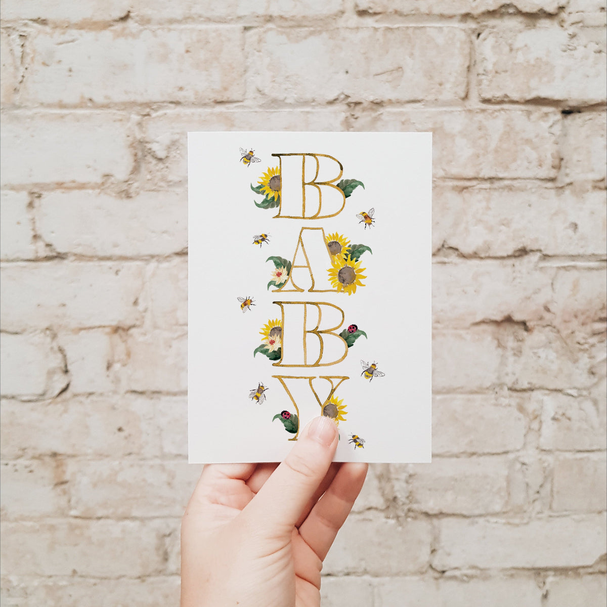 Hand holding a baby greeting card with sunflowers, bees, leaves and lady beetles against a brick wall