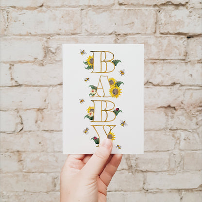 Hand holding a baby greeting card with sunflowers, bees, leaves and lady beetles against a brick wall