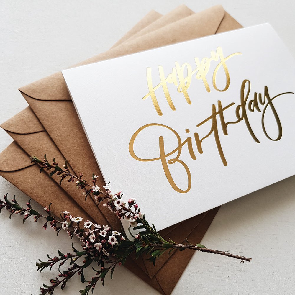 Gold foiled hand lettered happy birthday card ontop of brown envelopes with flowers