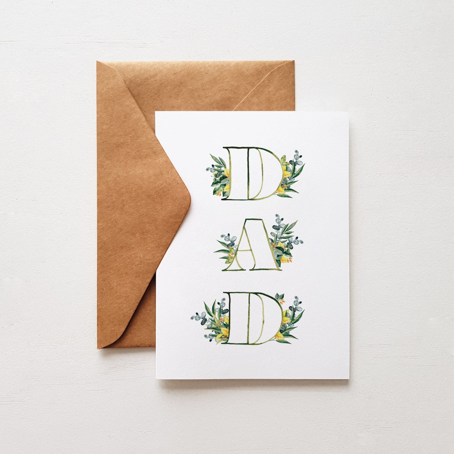 A greeting card with the word 'DAD' surrounded by leaf designs, folded with a brown envelope on a white background