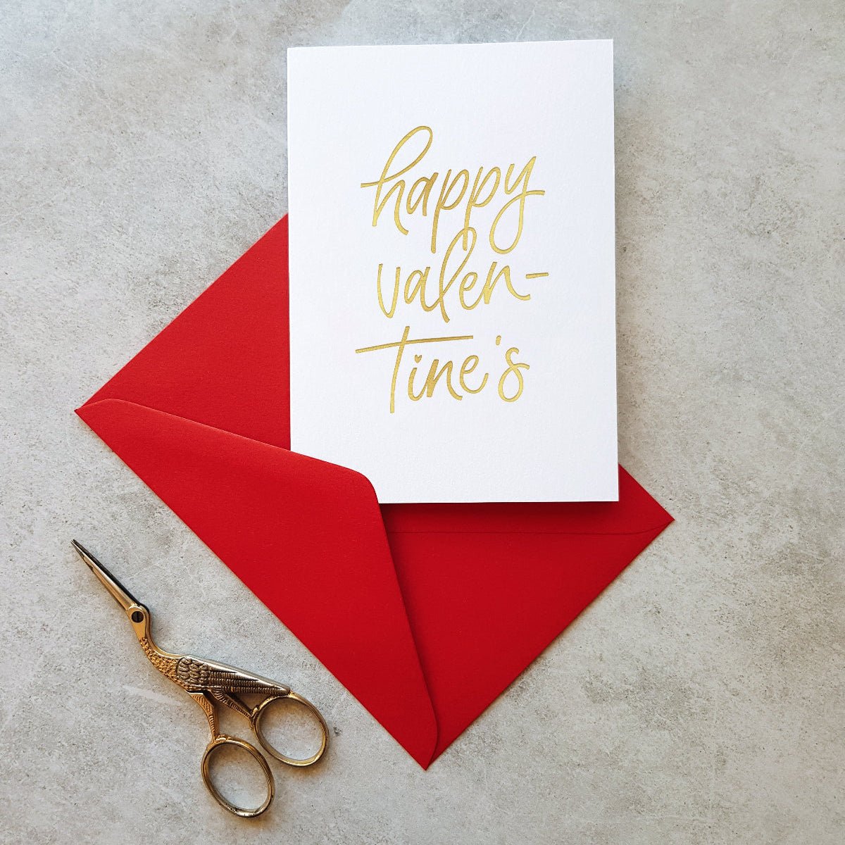 A white greeting card with gold foil script reading 'Happy Valentine's Day' on a red envelope, with gold scissors on a grey background.