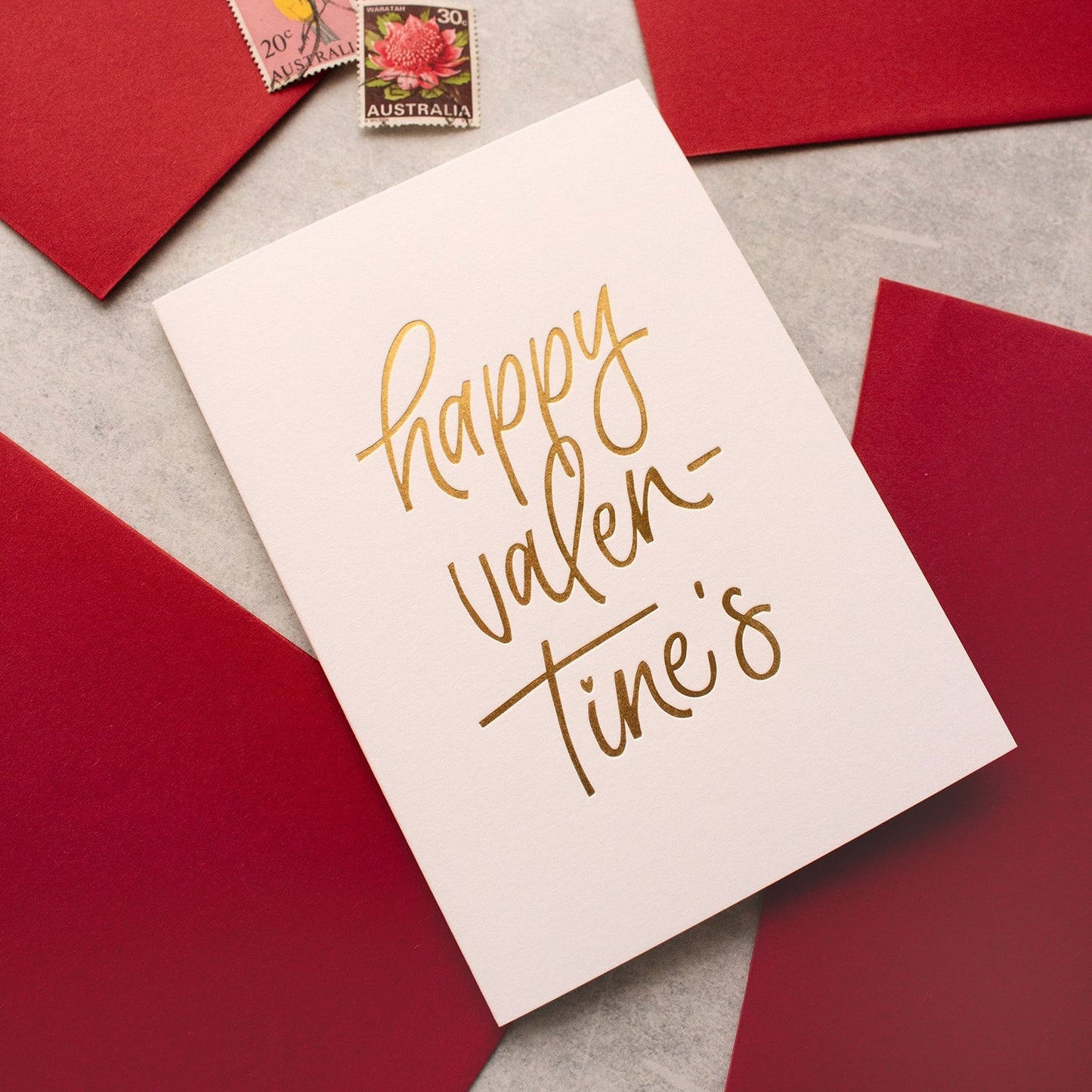 Valentine's Day greeting card with gold foil script 'happy valentine's' is arranged on a light grey surface with red envelopes