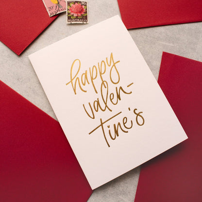 Valentine's Day greeting card with gold foil script 'happy valentine's' is arranged on a light grey surface with red envelopes