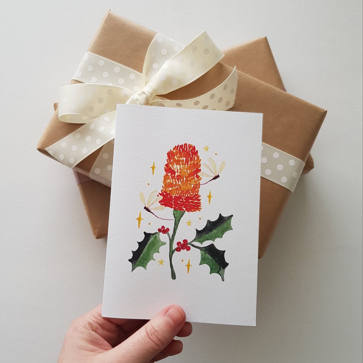 Hand holding a Christmas card featuring a banksia and holly illustration, ontop ofa stack of brown paper gift boxes tied with cream ribbon.