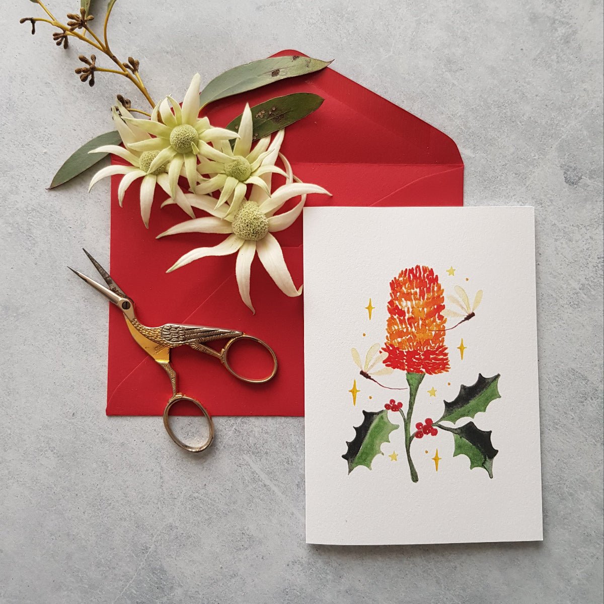 Red envelope with flowers, scissors and a greeting card with a red banksia, holly leaves and dragonflies