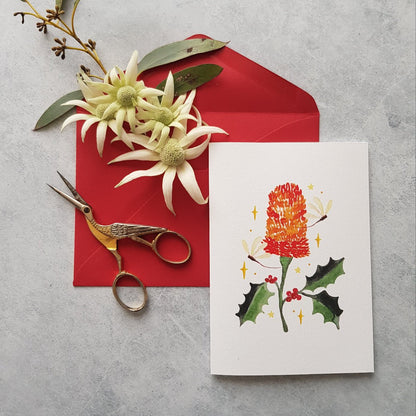 Red envelope with flowers, scissors and a greeting card with a red banksia, holly leaves and dragonflies