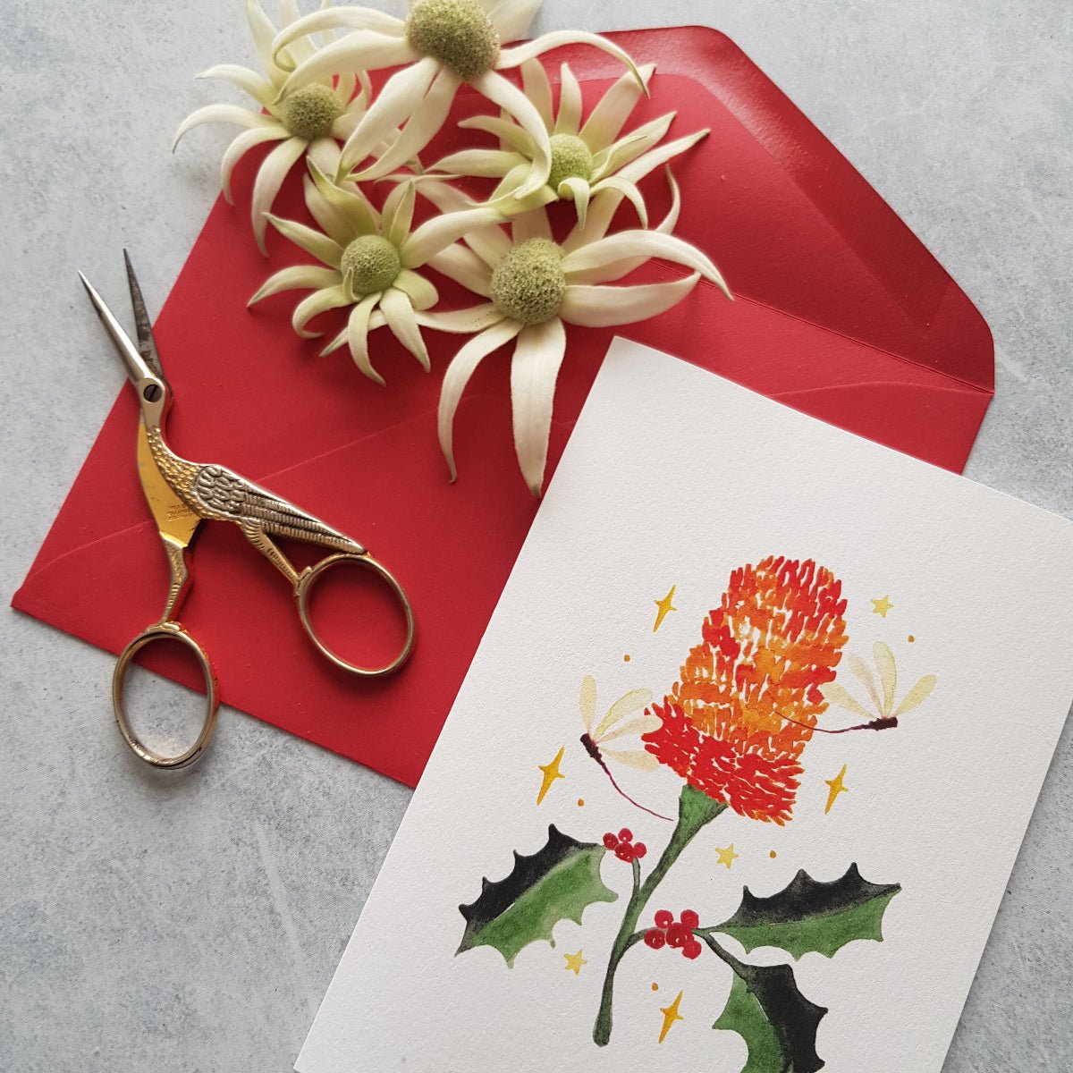 A white greeting card with a colorful illustration of holly leaves, red berries, banksia with a red envelope, gold bird scissors, and white flowers on a grey surface.