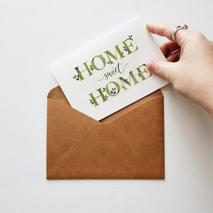 A hand holds a white card that reads 'HOME sweet HOME' in green decorative lettering above an open brown kraft paper envelope on a light background.