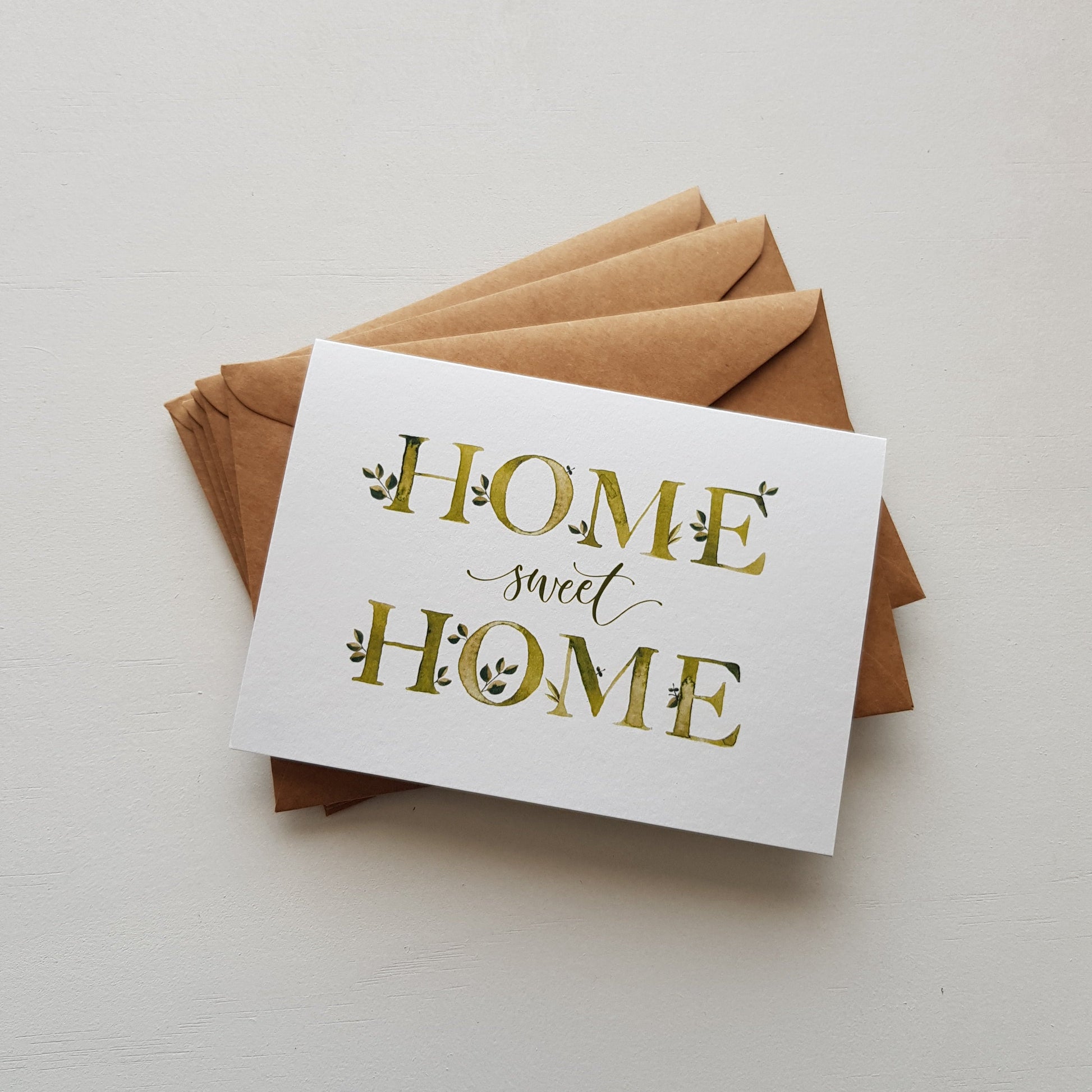 A white greeting card with the words 'HOME sweet HOME' printed in gold and dark green lettering, resting on top of a stack of brown kraft paper envelopes against a clean white background.