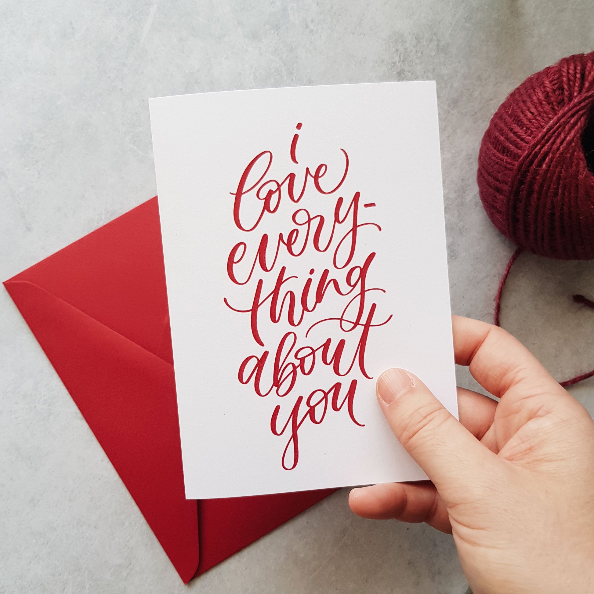 Hand holding a greeting card with red text 'I love everything about you' on a red envelope, with a ball of burgundy yarn.