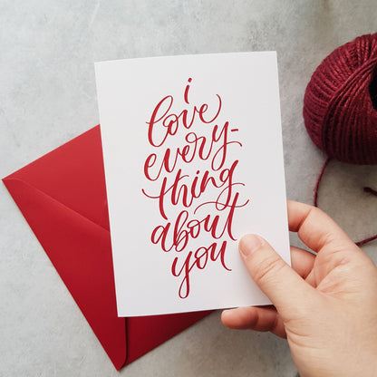 Hand holding a greeting card with red text 'I love everything about you' on a red envelope, with a ball of burgundy yarn.