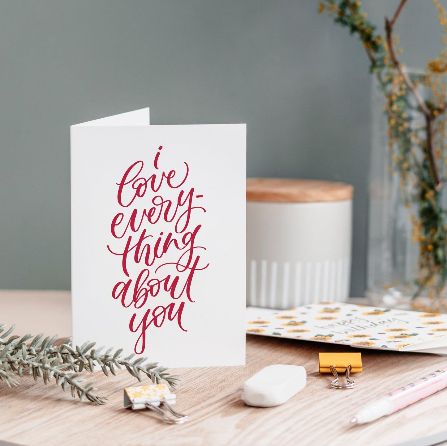 A white greeting card with elegant red cursive text reading 'love everything about you' stands open on a wooden tabletop. 