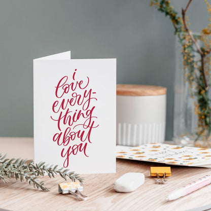 A white greeting card with elegant red cursive text reading 'love everything about you' stands open on a wooden tabletop. 