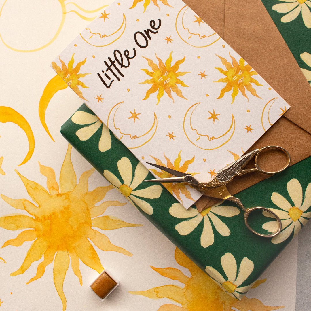 'Little One' baby greeting card with suns, moons, and stars, placed on a kraft envelope and daisy-patterned wrapping paper.