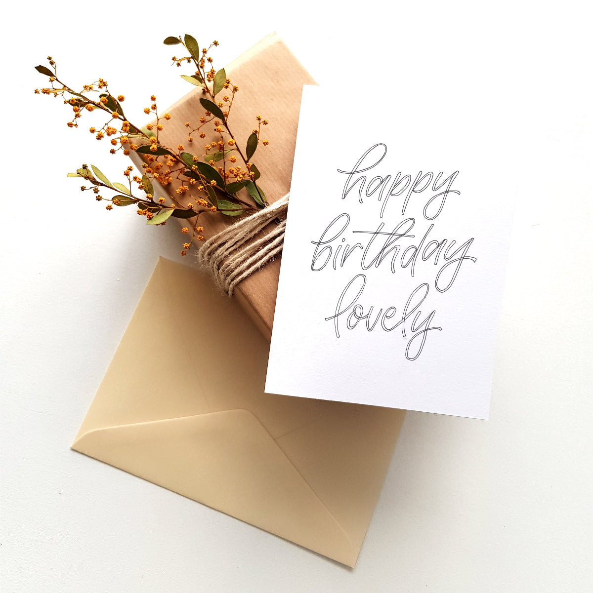 Greeting card 'happy birthday lovely' with dark grey script ontop of a wrapped present with flowers and cream envelope