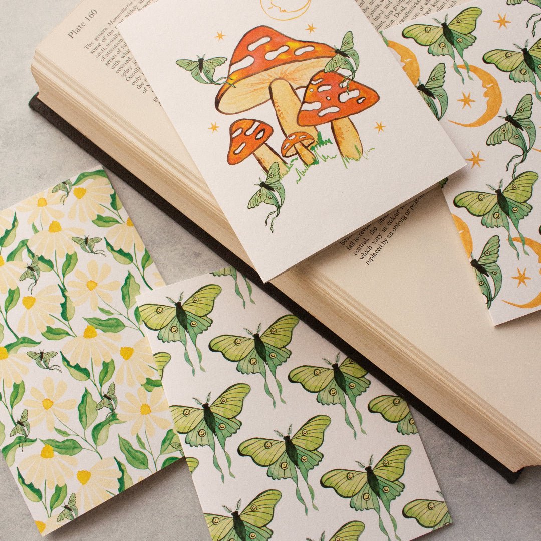 Collection of everyday cards featuring luna moths, daisies, orange-red mushrooms, yellow moons, and stars, on a book.